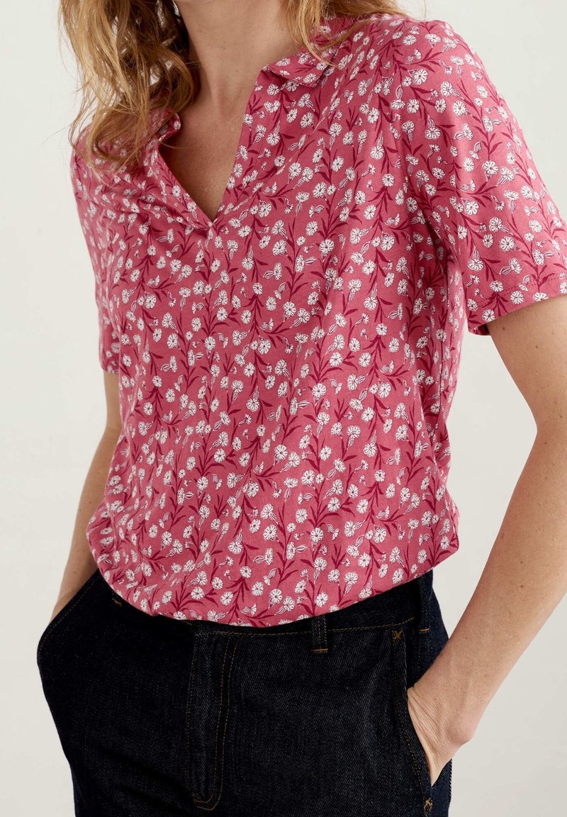 Femme portant un chemisier rose à manches courtes avec imprimé floral blanc, rentré dans un jean bleu foncé, mains dans les poches, sur fond uni.