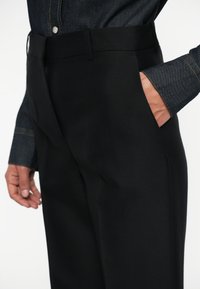 Zwarte op maat gemaakte broek met een gladde textuur, met zijzakken en een platte tailleband. Weergegeven naast een spijkerhemd.