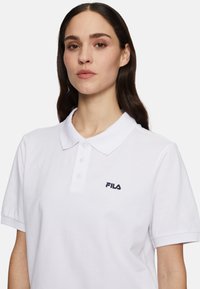 Vit polo-tröja i bomull, med klassisk krage, tre-knapps slits och en marinblå FILA-logotyp på vänster bröst.