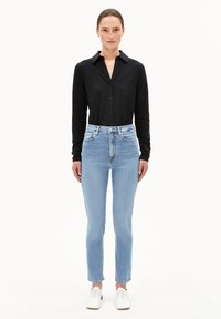 Vrouw in een zwarte lange mouwen met knopen shirt, lichtblauwe high-waisted jeans en witte veterschoenen, staand tegen een effen witte achtergrond.