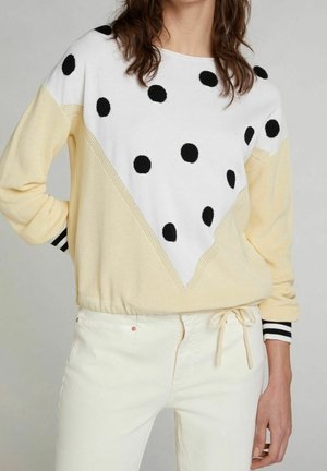 Kvinde iført en cremefarvet sweater med sorte prikker på en hvid trekantssektion og hvide bukser med snøre i taljen.