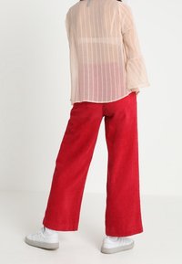 Transparente, hellfarbene Bluse mit gestreiftem Design, kombiniert mit weit geschnittenen, gerippten roten Hosen und weißen Sneakers, von hinten betrachtet.
