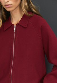 Veste zippée bourgogne avec un col, arborant une texture lisse et un zip argenté sur le devant.