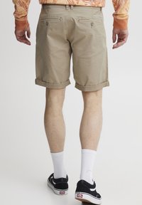 Pantalones cortos enrollados beige de algodón, con bolsillos traseros con botones, combinados con zapatillas negras y calcetines blancos sobre un fondo neutro.