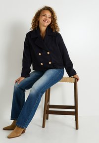 Veste bleue marine croisée avec des boutons dorés et des détails à côtes verticales, associée à un jean bleu délavé et des bottes chelsea beige, assis sur un tabouret.