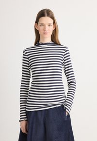 Langärmliges Oberteil in Marineblau und Weiß mit gestreiftem Muster, gerippter Textur und gewelltem Kragen. Kombiniert mit einem strukturierten Marineblauen Rock.