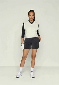 Mulher em pé com as mãos nos bolsos, vestindo um colete branco sem mangas sobre uma camisa de mangas longas preta, calções pretos, luva branca, meias e ténis.