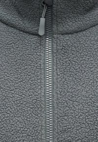 Κοντινό πλάνο γκρι υφάσματος φλις με μαύρο φερμουάρ που διαθέτει υφασμάτινο έλκτη με υφή στο κέντρο.