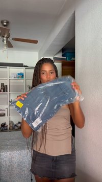 Zilas džinsu bikses caurspīdīgā iepakojumā, ar aizmugures kabatu un izbalējušu krāsu, ko tur cilvēks bežā blūzē un pelēkā svārkā.