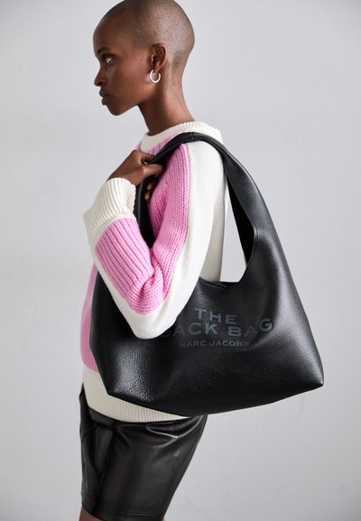 Mustast nahkainimese kott pehme tekstuuriga, millel on printitud tekst "THE SACK BAG MARC JACOBS." Mudel kannab roosa ja valge triibulist kampsunit.