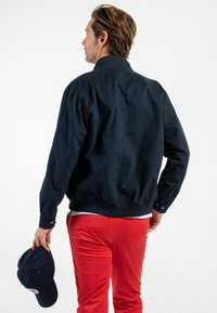 Veste bombardier bleu marine avec un col et des poignets élastiques, associée à un pantalon rouge. La personne tient une casquette bleu foncé dans une main.