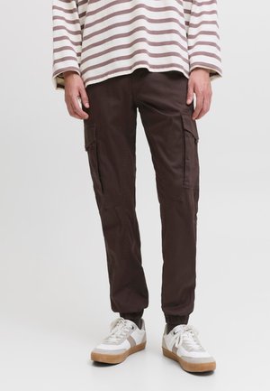 Personne portant un pantalon cargo marron foncé, des baskets blanches à semelles en gomme et une chemise blanche à manches longues avec des rayures horizontales marron.