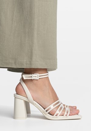 Białe sandały na obcasie z lakierowanej skóry, z szerokim paskiem, kwadratowym noskiem i klamrą przy kostce. Połyskujące wykończenie i masywny słupek na obcasie.