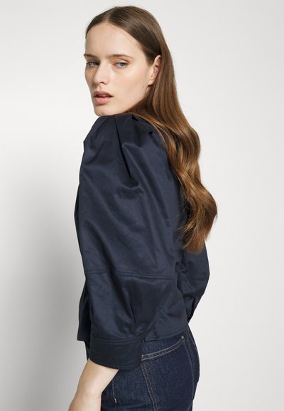 Ναυτικό μπλε, ελαφρύ μπουφάν με φουσκωτά μανίκια, ραμμένη εφαρμογή, διακριτική γυαλάδα και cropped μήκος. Υφασμάτινη υφή ορατή στις ραφές.