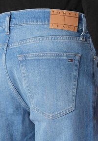 Jean en denim bleu clair avec une étiquette en cuir portant la mention "TOMMY JEANS". Comprend deux poches arrière et des détails de couture classiques.