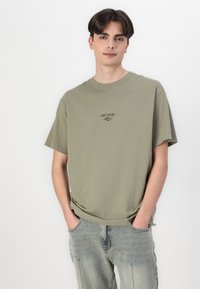 Olive green cotton t-shirt with a crew neck, short sleeves, and a small "Rip Curl" logo printed on the chest. Worn with light wash jeans.