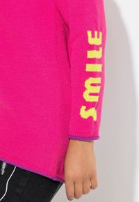 Heller pinkfarbener Pullover mit langen Ärmeln und gelbem "SMILE"-Text am Ärmel sowie lila Besatz an den Bündchen und dem Saum, kombiniert mit schwarzen Hosen.