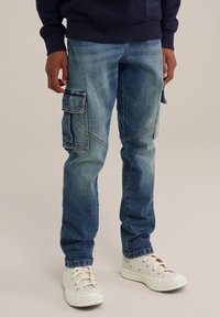 Pantalones cargo de denim azul claro con múltiples bolsillos laterales, un ajuste ceñido y áreas descoloridas, acompañados de zapatillas blancas.