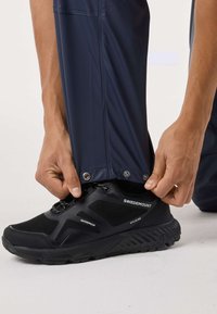 Musta vedenpitävä lenkkari, jossa on teksturoitu verkkomateriaali ja kumipohja. Henkilö säätää laivastonsinisten vedenpitävien housujen resoria, joissa on painonappikiinnitys.