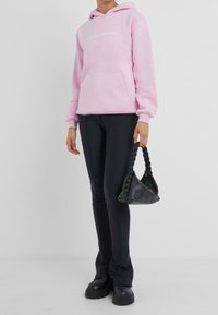 Felpa con cappuccio rosa chiaro con tasca frontale e testo bianco, abbinata a pantaloni aderenti neri e una borsa nera strutturata con manico intrecciato.