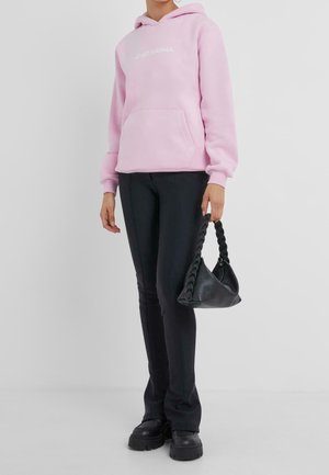 Felpa con cappuccio rosa chiaro con tasca frontale e testo bianco, abbinata a pantaloni aderenti neri e una borsa nera strutturata con manico intrecciato.