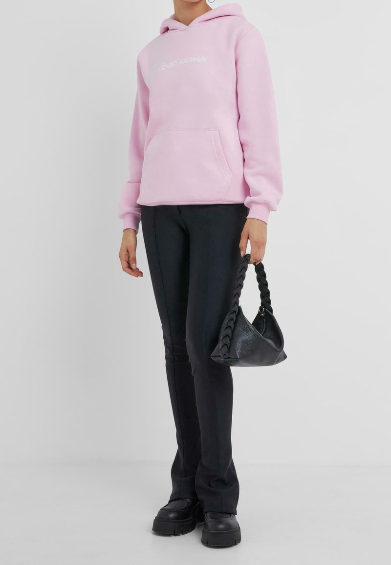 Felpa con cappuccio rosa chiaro con tasca frontale e testo bianco, abbinata a pantaloni aderenti neri e una borsa nera strutturata con manico intrecciato.