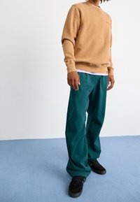 Felpa beige con collo a coste; pantaloni larghi turchesi con polsini elastici; indossati con sneakers nere su una superficie azzurro chiaro.