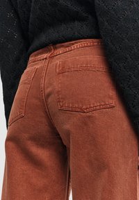 Roestkleurige spijkerbroek van denim met een gestructureerde afwerking. Voorzien van twee achterzakken en een medium taille. G gedragen met een gestructureerd zwart top.