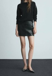 Maglione nero lavorato a maglia con polsini a coste, abbinato a una mini gonna in pelle nera. L'outfit include décolleté nere con punta appuntita.