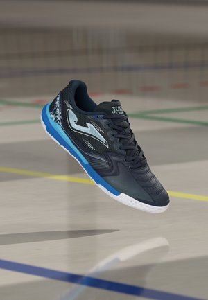 Zwart-blauwe sportschoen voor binnen met wit logo en veters, zwevend boven een gymzaalvloer met kleurrijke veldlijnen.