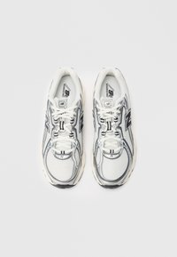Une paire de chaussures de course New Balance blanches et argentées avec une tige en mesh, des détails noirs et des lacets blancs sur un fond blanc uni.