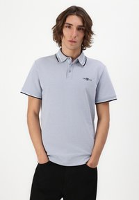 Lys grå polo t-shirt lavet af struktureret stof, med en ribbet krave med kontrasterende sorte striber og et lille logo nær brystet.