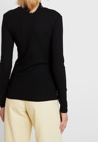 Mustaa pitkähihainen ribbitekoinen paita, jossa on sopiva muotoilu. Takana näkyy pyöreä kaula-aukko ja sileä pinta. Yhdistetty vaaleansävyisiin housuihin.