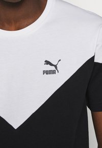 Černobílé tričko Puma nošené osobou, na kterém je na bílém středním dílu na hrudi s diagonálním rozdělením barev uprostřed zobrazeno logo Puma.