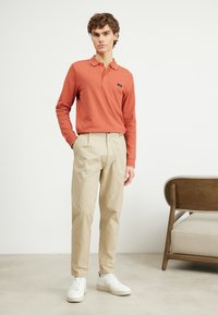 Polo a maniche lunghe corallo con colletto bianco, abbinato a pantaloni beige e sneaker bianche. Il modello è in piedi su uno sfondo semplice.