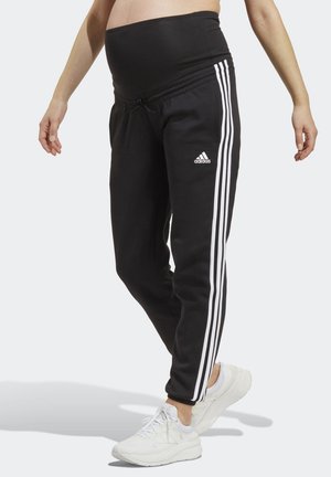 Persona embarazada vistiendo pantalones deportivos de maternidad negros de Adidas con rayas blancas, combinados con zapatillas blancas sobre un fondo claro.