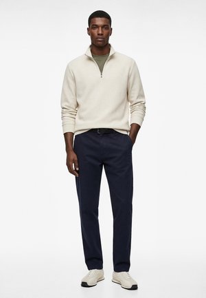 Mann trägt einen beigefarbenen Pullover mit Viertel-Reißverschluss, olivgrünes Hemd, marineblaue Hose und weiße Turnschuhe, steht mit einer Hand in der Tasche.