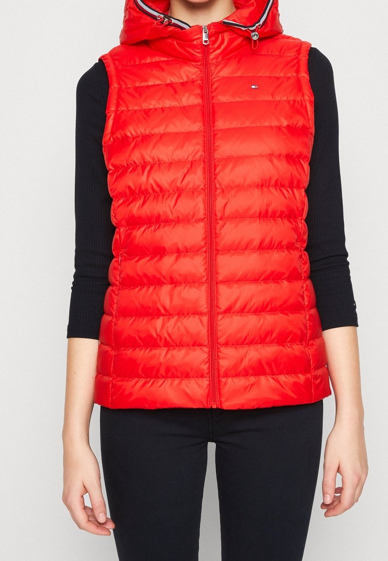 Červená prošívaná vesta na zip vpředu, s připojenou kapucí a šitími detaily. Vyrobená z lesklého, izolovaného materiálu. Nositelná přes černé tričko s dlouhým rukávem.