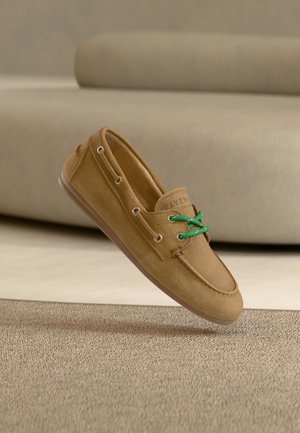 Lichtbruine suède loafer met groene veters, rustend op een beige tapijt voor een bijpassende beige bank.