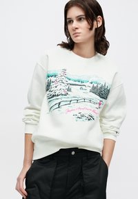 Sweatshirt branco com uma estampa de cena de inverno, incluindo uma árvore coberta de neve, montanhas e um esquiador, destacado por detalhes em rosa e verde-água.