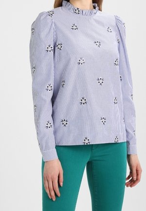 Vrouw die een blauw-wit gestreepte blouse met lange mouwen en bloemenprints draagt, gecombineerd met getailleerde teal-kleurige broek, staand met ontspannen armen langs haar lichaam.