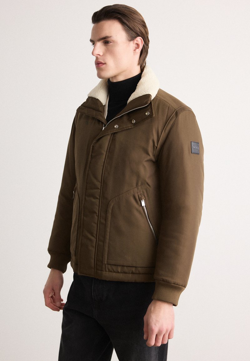 Brun polstret jakke med sherpa-krave, lynlåslommer, elastiske manchetter og et logo-patch på ærmet. Glat tekstur, alsidigt design.
