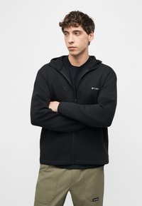 Νέος άνδρας με καστανά σγουρά μαλλιά, φορώντας μαύρο φούτερ με φερμουάρ και χακί παντελόνι, στέκεται με τα χέρια σταυρωμένα και κοιτάζει πλάγια.