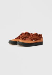 Brūni zamša sporta apavi ar zemu augstumu, ar melniem gumijas zolemi, auklu aiztaisīšanu un baltu Nike swoosh sānos.