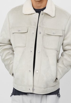 Homme portant une veste en peau retournée beige clair avec des boutons pression et des poches à rabat sur la poitrine, pull noir en dessous, mains dans les poches.