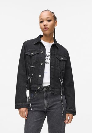 Schwarze Jeansjacke mit einem verkürzten Design, ausgestattet mit einer Knopfleiste vorne, Brusttaschen und Kordelzügen am Saum. Kombiniert mit schwarzen Jeans.