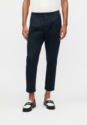 Mężczyzna noszący krótkie czarne spodnie z guzikiem i zamkiem błyskawicznym, w czarno-białych mokasynach, stojący na jednolitym tle.