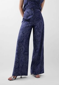 Pantalons à jambe large en tissu bleu marine avec un motif texturé ressemblant à des vagues. Présentent une taille haute et un détail de bouton argenté.