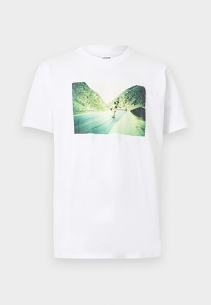 Wit T-shirt met een afgedrukte foto van een skateboarder die een bergweg afdaalt, omgeven door groene rotsachtige heuvels onder een heldere hemel.