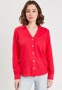Rotes Leinen-Hemd mit Knopfleiste, Kragen, langen Ärmeln, einer Brusttasche und weißen Knöpfen. Sanfte Textur und lockere Passform.